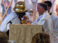 Una imagen de una ceremonia religiosa, posiblemente un servicio católico, con un hombre en traje blanco y corbata, sosteniendo una copa de vino. En el fondo, se ve a un hombre con un sombrero dorado y una figura religiosa en la pared. La escena parece ser en un templo o iglesia, con una estructura de madera y detalles artísticos.