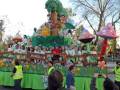 Un carnaval con una gran carroza verde y amarilla, decorada con flores y animales. La carroza está flanqueada por dos grandes setas de colores rosa y azul. La carroza está lleno de personas vestidas con trajes de personajes de dibujos animados. La carroza está rodeada por un público que observa la carroza con interés.