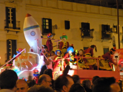 Un globo gigante en forma de cohete blanco y rojo iluminado, con un personaje de dibujos animados en la parte superior. La imagen se toma durante una celebración nocturna, con gente observando desde el suelo. La escena está ambientada en una calle con edificios blancos y ventanas negras.