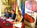 A tres personas sentadas en una mesa con documentos y banderas, incluyendo la de España y la UE. A la derecha se encuentra un cartel promocional para SIMOF 2013, que se celebra del 31 de enero al 5 de febrero.