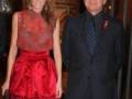 Una pareja elegante posa juntos en un evento formal, ella con un vestido rojo y él en traje negro con corbata de lunares.
