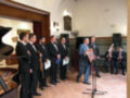 Un grupo de músicos en trajes formales se prepara para un concierto, con un piano y otros instrumentos al fondo.
