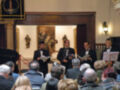 Un grupo de músicos en trajes formales se encuentra frente a un público en una sala decorada con arte religioso. La imagen muestra a los músicos leyendo partituras mientras el público escucha atentamente.