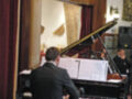 Un hombre en traje negro toca un piano de cola en una sala con paredes blancas y marrones. La imagen muestra la parte trasera del piano, el teclado y las partituras en blanco. Hay una columna de mármol y un cuadro colgando en la pared al fondo.