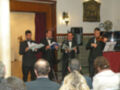 Un grupo de músicos en trajes formales se presentan en un salón con una audiencia. El ambiente es elegante, con cortinas rojas y un reloj en la pared. Los músicos tocan instrumentos de cuerda y viento, mientras el público observa atentamente.