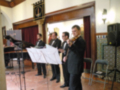 Un grupo de músicos en trajes formales se preparan para tocar música clásica. La imagen muestra a cuatro hombres, dos de ellos tocando flautas y el otro un piano. La escena se desarrolla en una sala con decoración tradicional, incluyendo un cartel y una lámpara de pared. La atención se centra en la concentración y el arte musical que están realizando.
