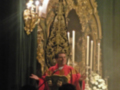 Un sacerdote en vestido rojo y dorado frente a una imagen sagrada, con velas encendidas en la parte superior.