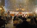 Interior de una iglesia con un altar central, vitrales y estatuas. Personas sentadas en filas de sillas, algunos levantando las manos. Altar con cruz y estatuas religiosas. Ambiente sereno, iluminado por lámparas de techo y vitrales.