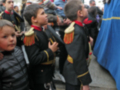 Un grupo de niños vestidos con uniformes militares, algunos con sombreros y otros con campanas en las manos, están participando en una procesión o ceremonia. El fondo muestra un edificio con una pared de ladrillo y una bandera azul ondeando.