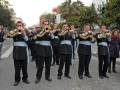 Un grupo de músicos militares toca instrumentos de viento en una calle con árboles frutales y edificios a lo lejos.
