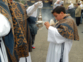 Un niño en una procesión religiosa, vestido con un hábito blanco y azul ornado, sostiene una antorcha encendida. Al fondo, una figura en un trono y personas vestidas con hábitos religiosos.