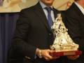 Un hombre en traje sostiene un premio de plata, con una escultura religiosa. Detrás, se ve un mural y otro hombre en traje.