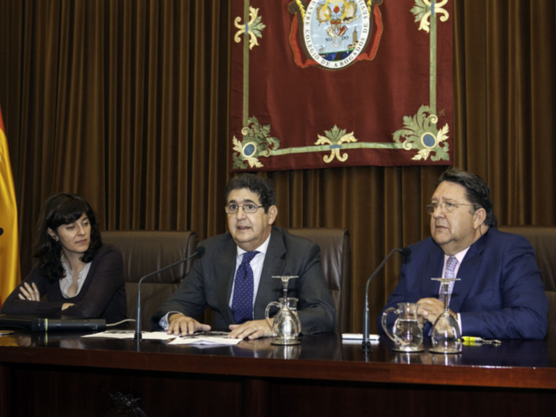 Tres personas sentadas en una mesa con un escudo y una bandera en el fondo.
