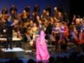 Una cantante en un vestido rosa brillante en el escenario principal, con un orquesta clásica detrás, incluyendo violines y violonchelos, en una representación de ópera o concierto.