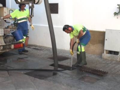 Trabajadores de mantenimiento en una calle, uno aplica pintura y otro usa un cepillo para limpiar el asfalto.