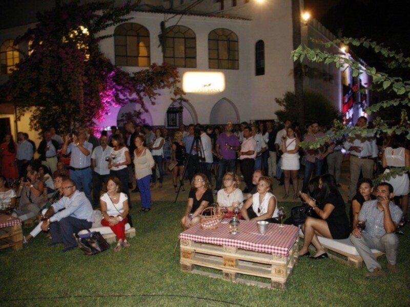 Una multitud de personas disfruta de una fiesta nocturna en un jardín con mesas de madera y sillas, adornadas con mantel rojo y blanco. La iluminación es cálida, destacando la arquitectura de un edificio blanco con ventanas y una gran lámpara central. La atmósfera es festiva, con gente conversando y bebiendo en un ambiente relajado.