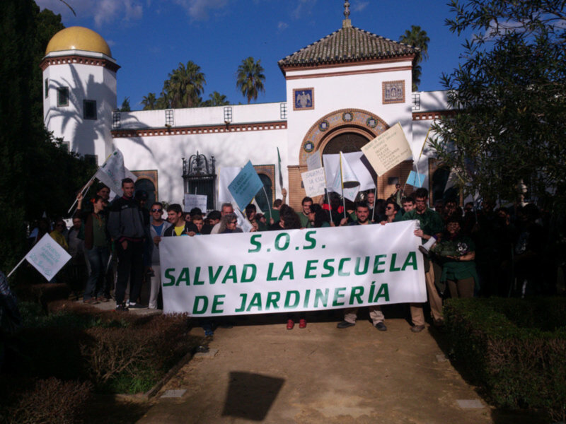 Un grupo de personas sostiene un cartel blanco con la leyenda "S.O.S. Salvad la Escuela de Jardinería" frente a un edificio blanco con una cúpula amarilla y un torreón. El cielo es azul claro con algunas nubes dispersas, y hay palmeras al fondo. La imagen parece ser de una manifestación o protesta por la protección de un establecimiento educativo.