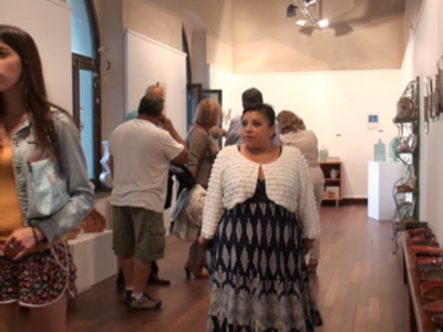 Una mujer con vestido azul y blanca camina por un pasillo de exposiciones, mientras otros visitantes observan arte en el fondo. La atmósfera es cultural y educativa, con una combinación de arte contemporáneo y objetos decorativos.