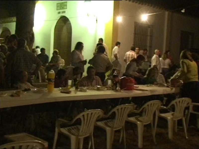 Una noche de fiesta en una terraza con gente reunida alrededor de mesas y sillas blancas. La iluminación es natural, proveniente de luces colgantes y una luz en la pared. La atmósfera es festiva, con muchos participantes disfrutando de la noche.