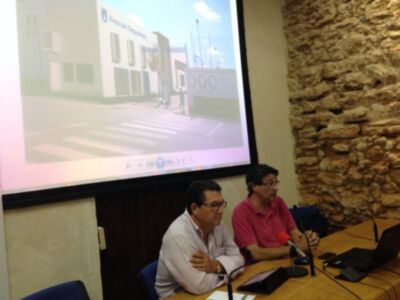 Dos hombres sentados en una sala de conferencias, uno frente a un proyector que muestra imágenes de la ciudad. La pared del fondo es de ladrillo y hay una mesa con portátiles, auriculares y cables. La imagen en el proyector muestra una calle con un edificio de oficinas y una señal de tráfico.