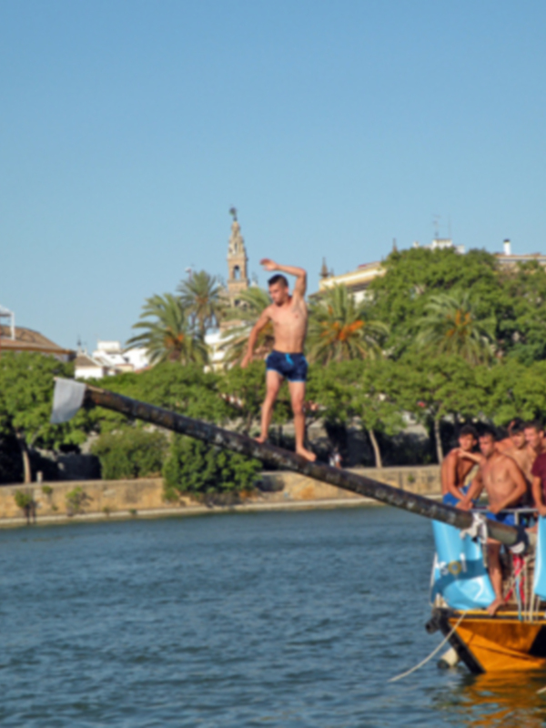 Un joven se prepara para saltar de un bote amarrado en el agua, rodeado por otros jóvenes y palmeras. En la distancia se ve una torre histórica con un faro.