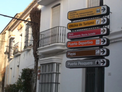 Se pueden observar varios carteles de dirección en un edificio blanco con balcones y ventanas. Los carteles indican diferentes destinos como Puerto Pesquero, Puerto Deportivo, Playas y Oficina de Turismo. También se puede ver un poste con una lámpara en la parte superior izquierda de la imagen.