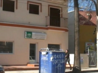 Un edificio de dos plantas con ventanas y balcones. En la parte superior, hay un cartel verde con letras blancas que parece ser el nombre de una empresa. En la calle, hay un contenedor azul con letras blancas y un coche blanco en el fondo.