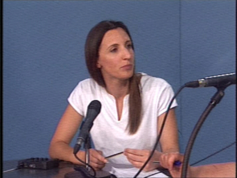 Persona hablando en un evento con micrófono y material de presentación en la mesa.