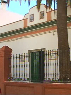 Fachada de una casa colonial con detalles en verde y blanco, palmeras y un coche azul aparcado.