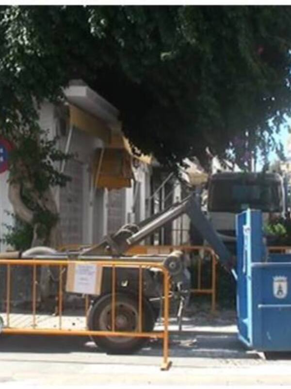 Trabajos de mantenimiento en una calle de la ciudad, con una máquina de limpieza y un contenedor azul en el camino.