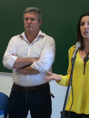 Una mujer en una camisa amarilla habla frente a un hombre con los brazos cruzados en una sala de conferencias con una pizarra verde y un portátil en el escritorio.