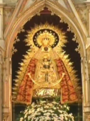 Imagen de una iglesia con un altar adornado, en el centro se encuentra una imagen de la Virgen María rodeada por flores y decoraciones doradas. El estilo arquitectónico es neogótico con detalles en madera y piedra.