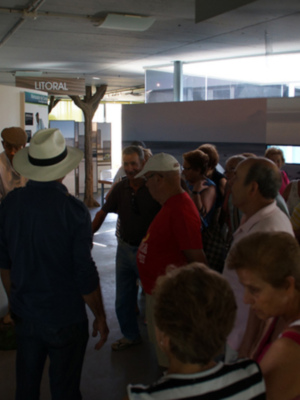 Grupo de personas en una sala de exposiciones, mirando un televisor colgado en la pared. La imagen muestra detalles de una sala con iluminación natural y un cartel que dice "LITORAL".