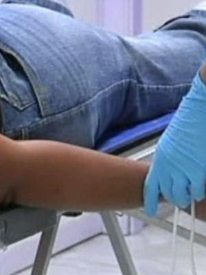 En un hospital, una persona se somete a una prueba médica. Se observa el brazo de la paciente extendido sobre un dispositivo médico, mientras que una mano con guantes azules aplica un tubo a la muñeca. La escena sugiere una exploración o análisis de sangre, probablemente para diagnóstico médico.
