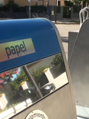 Puerta de papelera con la palabra 'papel' en español, en un entorno urbano con flores y coches a la distancia.
