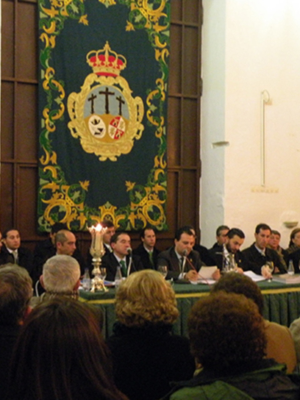 Conferencia en un salón con una bandera de la ciudad en el fondo. Los asistentes están sentados en mesas, algunas personas sonríen y otras escuchan. La iluminación es suave y la decoración incluye una bandera de la ciudad en el fondo.