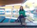 Una joven posa en la plataforma de un estadio deportivo, con una camiseta verde y zapatillas rojas. El fondo muestra una piscina de natación con líneas azules y amarillas, y varios espectadores en los stands.