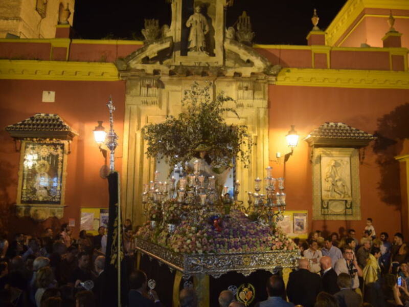 Fiesta religiosa con una imagen de la Virgen en una procesión nocturna, rodeada de flores y acompañada por una multitud de personas.