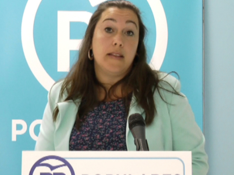 A una mujer con un fondo azul claro y blanco, posiblemente en un evento o conferencia. La mujer está vestida con una chaqueta blanca y tiene el cabello largo y ondulado. En la imagen, también se puede ver un logo que parece pertenecer a una organización o empresa, aunque no se puede leer claramente.
