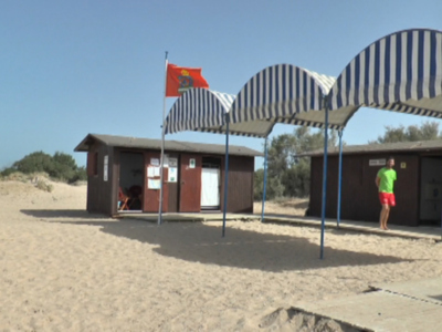 Una imagen de un pequeño edificio en la playa con toldos azules y blancos, una bandera roja ondeando, un hombre en shorts verdes caminando y arena blanca.