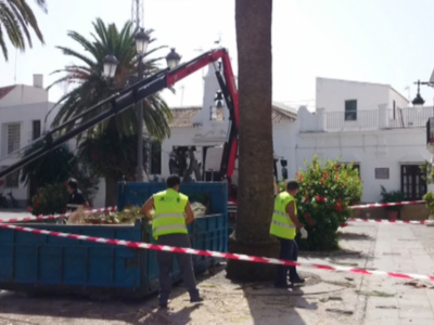 Trabajadores con chalecos amarillos y guantes trabajan en un contenedor azul cerca de un árbol. Hay una grúa roja y negra levantando objetos en el cielo, con palmeras y edificios blancos al fondo. La calle está bloqueada por cinta roja y blanca.