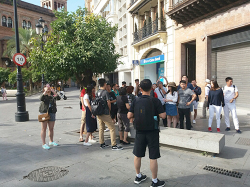 Un grupo de personas caminan por una calle con edificios históricos y árboles verdes. La imagen muestra a varias personas de diferentes edades, algunas con mochilas y otros con bolsos. La escena parece ser en un día soleado, con el cielo azul y la luz del sol brillando sobre los edificios.