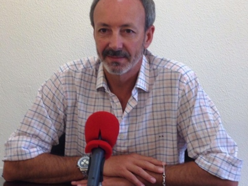 Un hombre con barba y bigote, vestido con una camisa de cuadros azules y blancos, se sienta frente a un micrófono rojo. La imagen sugiere una conferencia o entrevista informativa en un entorno formal.