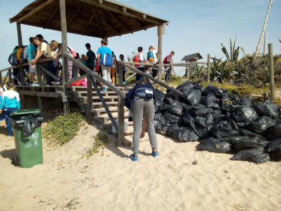 Una persona en ropa deportiva se prepara para recoger basura en una playa, con bolsas de plástico y un contenedor de residuos al lado. La escena muestra a varios individuos en la plataforma de madera, posiblemente supervisando el trabajo.