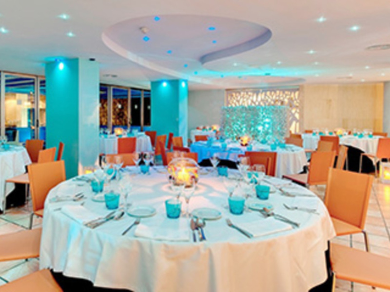 Salón de eventos con mesas decoradas con mantel blanco y azul, sillas naranjas, velas y decoración en azul claro.