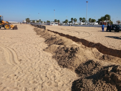 Una vista de la playa con un tractor amarillo trabajando en el suelo, mientras que varios vehículos y personas observan la obra. El cielo es claro y el paisaje está rodeado de árboles y edificios en la distancia.