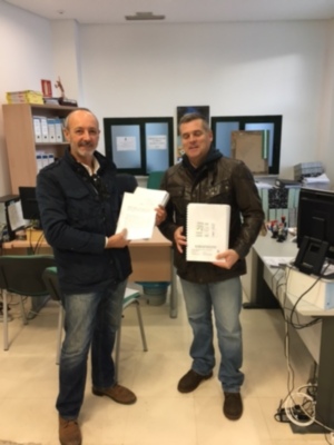 Dos hombres en un despacho, uno sosteniendo un documento mientras el otro lo observa. Ambos están en una oficina con estantes de libros y computadoras en el fondo.