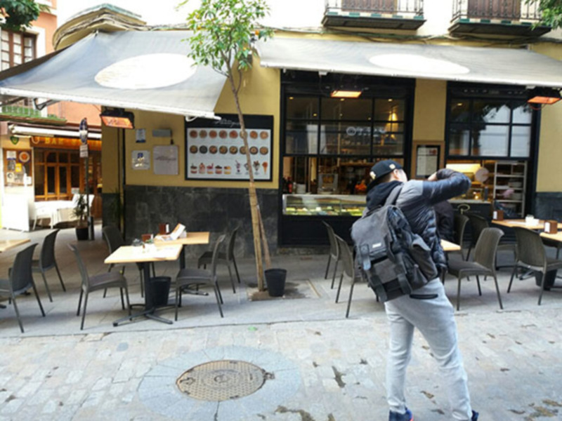 Persona caminando frente a un café con mesas al aire libre y una variedad de postres en la vitrina.