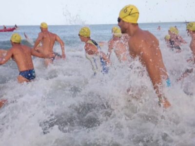 Una imagen de nadadores en la playa, con agua salobre y espuma. Los nadadores llevan gorras amarillas y trajes de baño azules, con una persona en un bote rojo a la izquierda. La escena captura el esfuerzo y la energía de los nadadores en un día soleado.