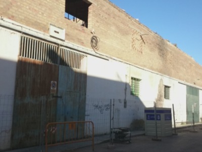 Fotografía de una pared de ladrillo con un hueco y un reloj en la parte superior. Hay una puerta de metal oxidada con barras de seguridad y un poste de luz en el lado izquierdo. El cielo es claro y azul.